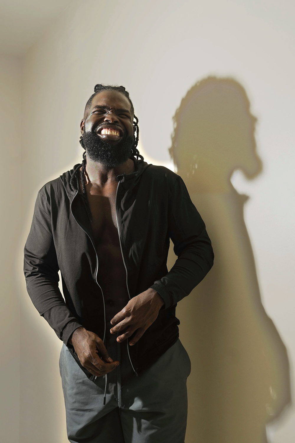 In a dark open-collar shirt, leaning on a pale wall mid-laugh, his shadow thrown large behind him.