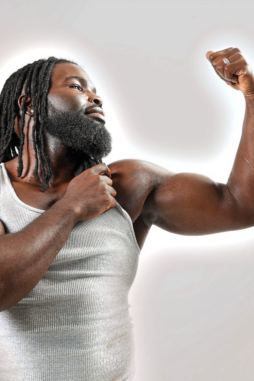 A white tank against a glowing wall, flexing one arm and pointing to it, eyes up.