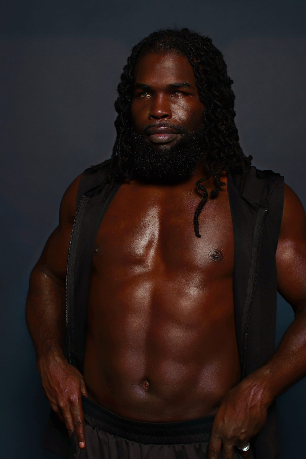 An open black vest over a bare, defined torso, squared to camera in a dark studio.