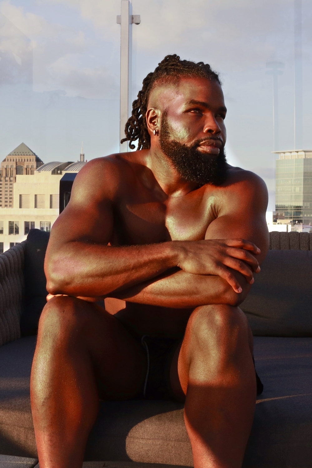 Seated on a rooftop at dusk, arms folded over one knee, the skyline behind him.