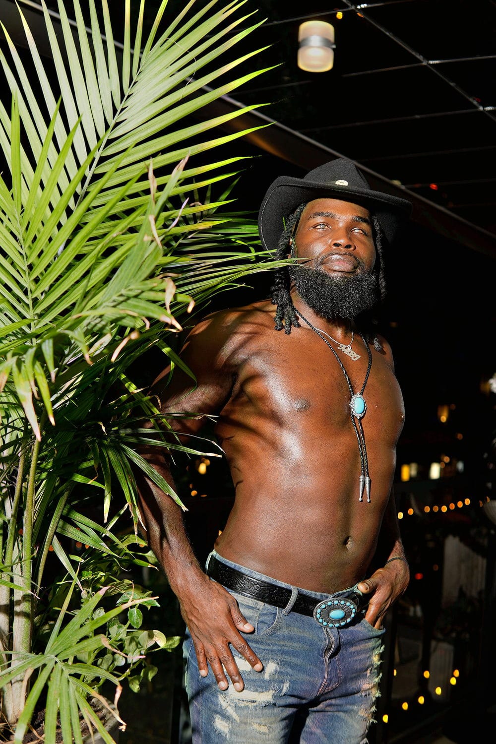 On a string-lit patio among palm fronds, in a cowboy hat and turquoise, hand on his belt.
