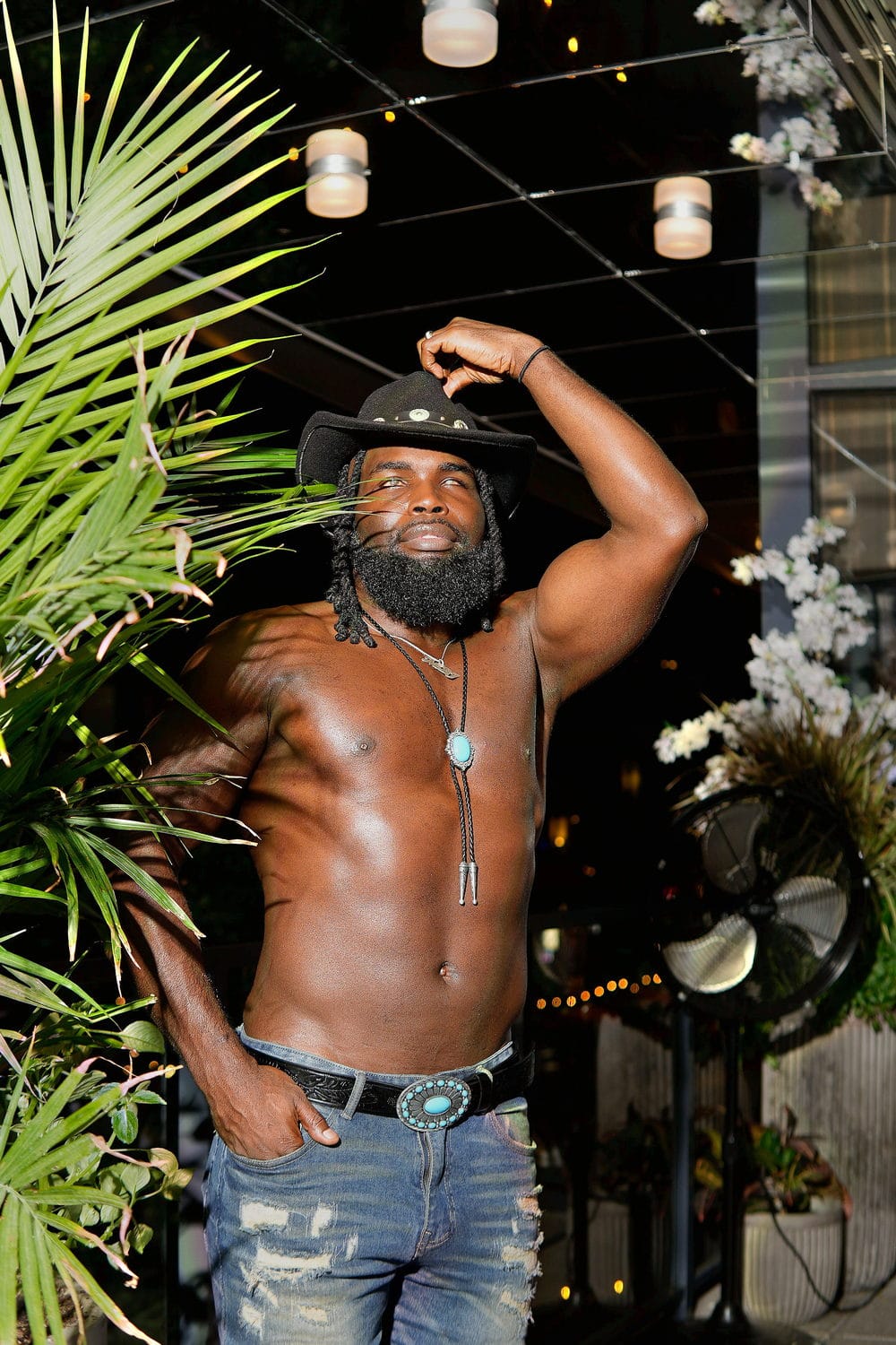 A cowboy hat tipped back on a night patio, arm raised to the brim, easy confidence.