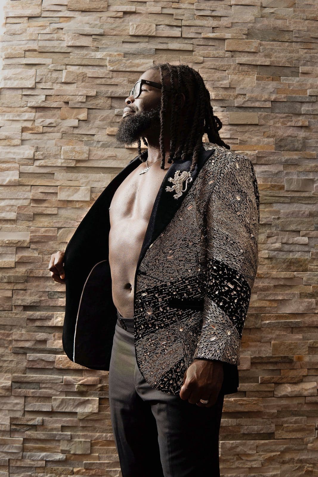 Stone wall, open silver sequin jacket, glasses, chin up.