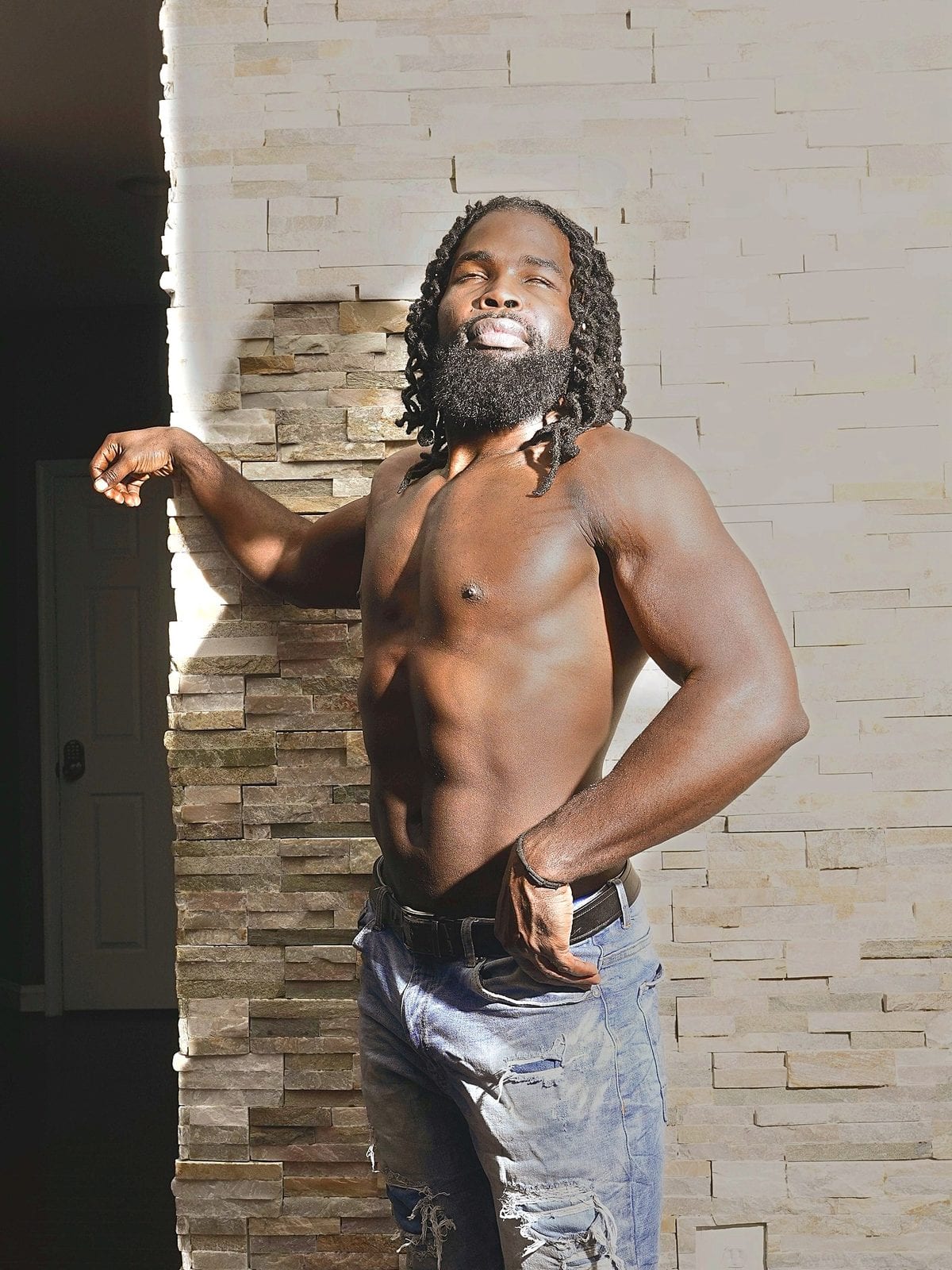 Stone wall, shirtless side flex, beard angled up, doorway light through the back of frame.