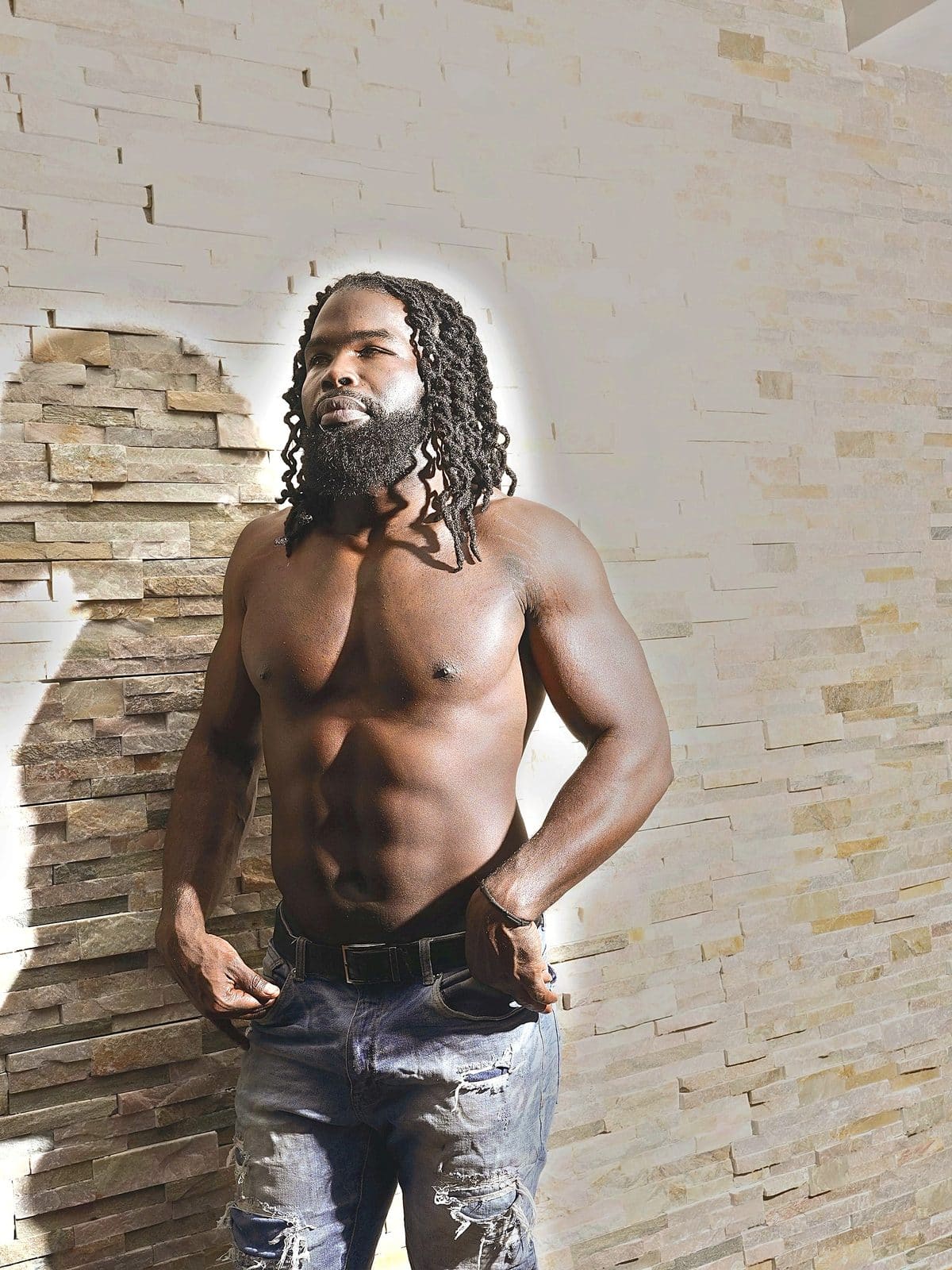 Stone wall, shirtless three-quarter view, hands at jeans, looking off-camera, considered.