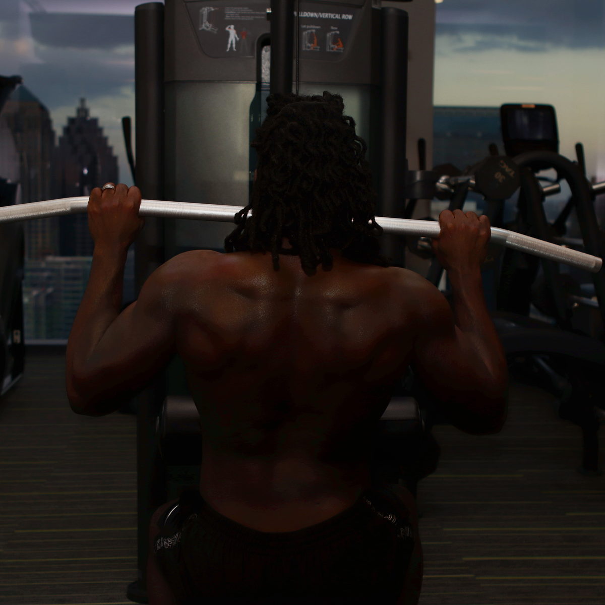 An overhead bar pull in a skyline gym, back to camera, the city through the windows.