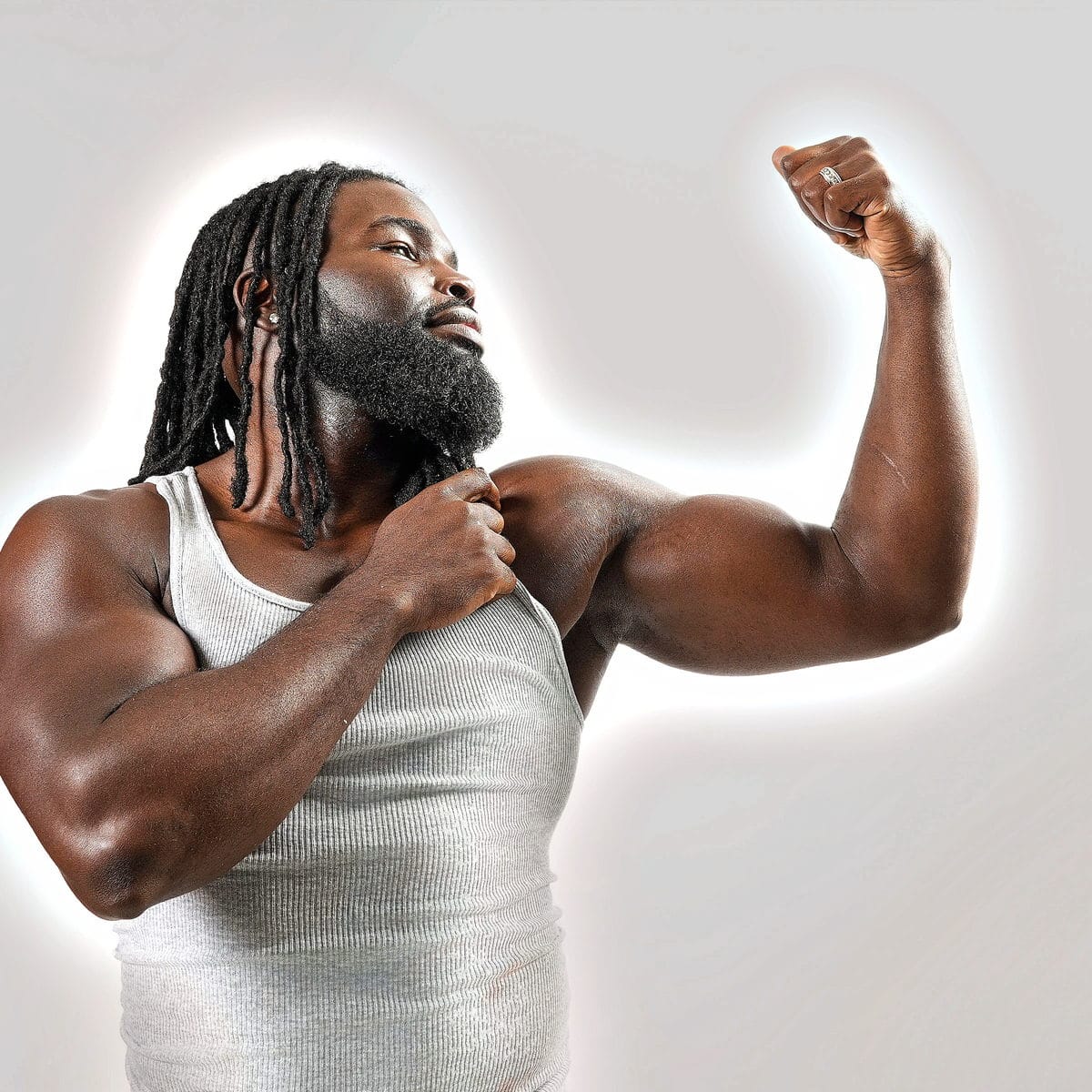 In a white tank against a bright wall, flexing one arm, eyes lifted.