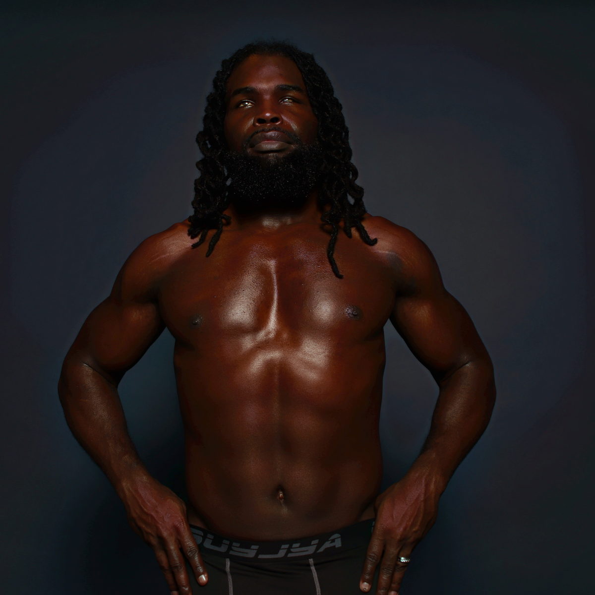 A shirtless frontal stance in a dark studio, hands at the waistband, defined midsection.