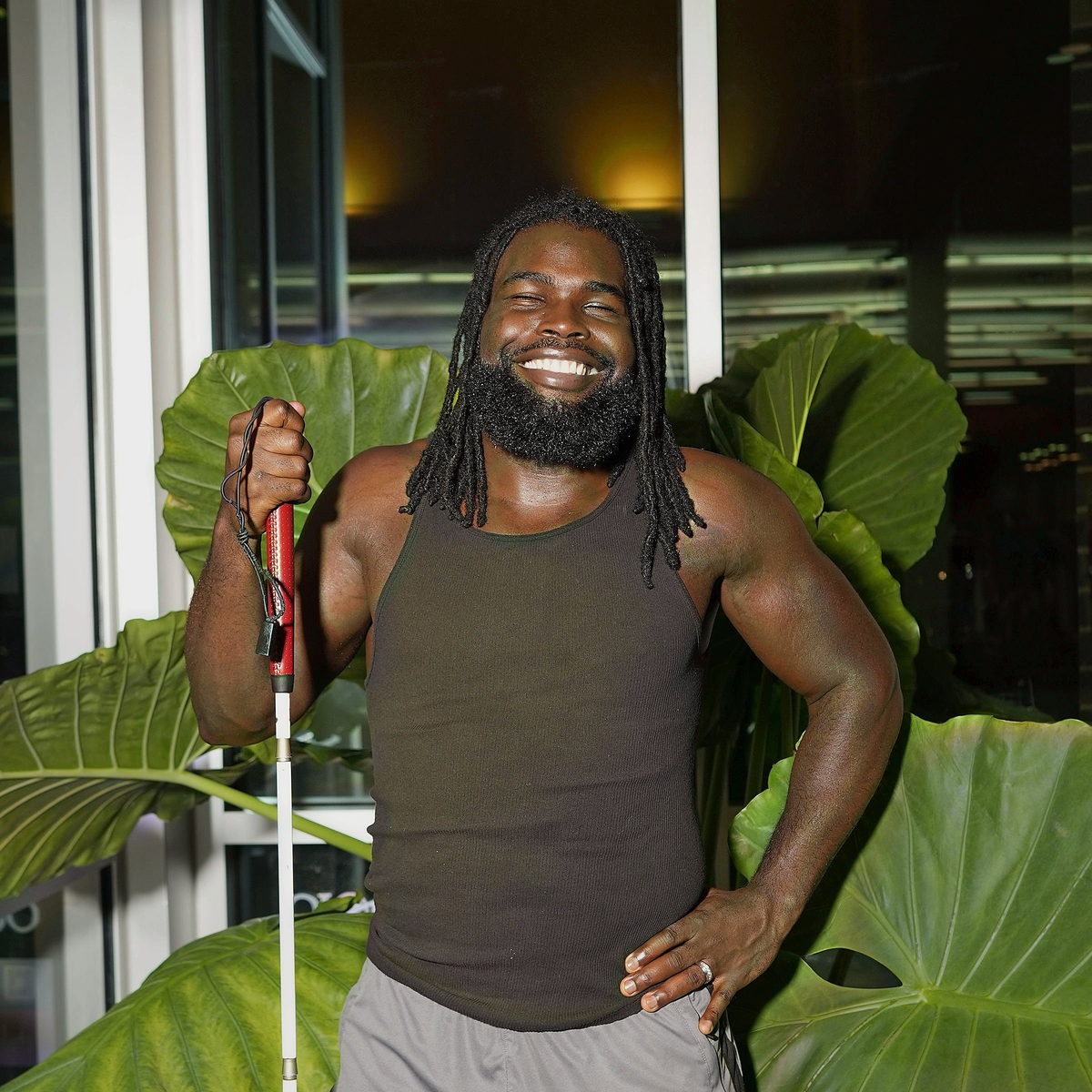 Among broad green leaves in an olive tank, a white cane in hand and a wide genuine smile.